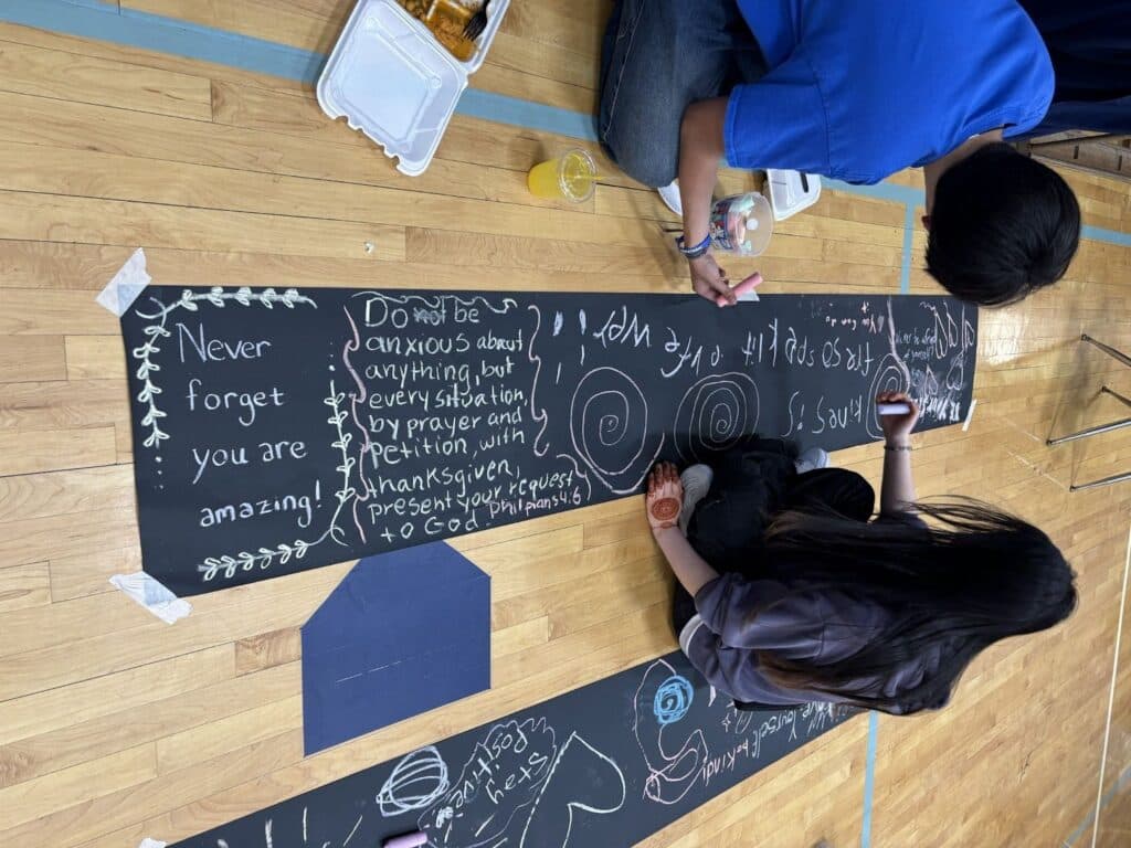 Two people sitting on a gym floor writing encouraging messages with chalk on long black paper. The messages include quotes about being amazing and not worrying, surrounded by decorative drawings.