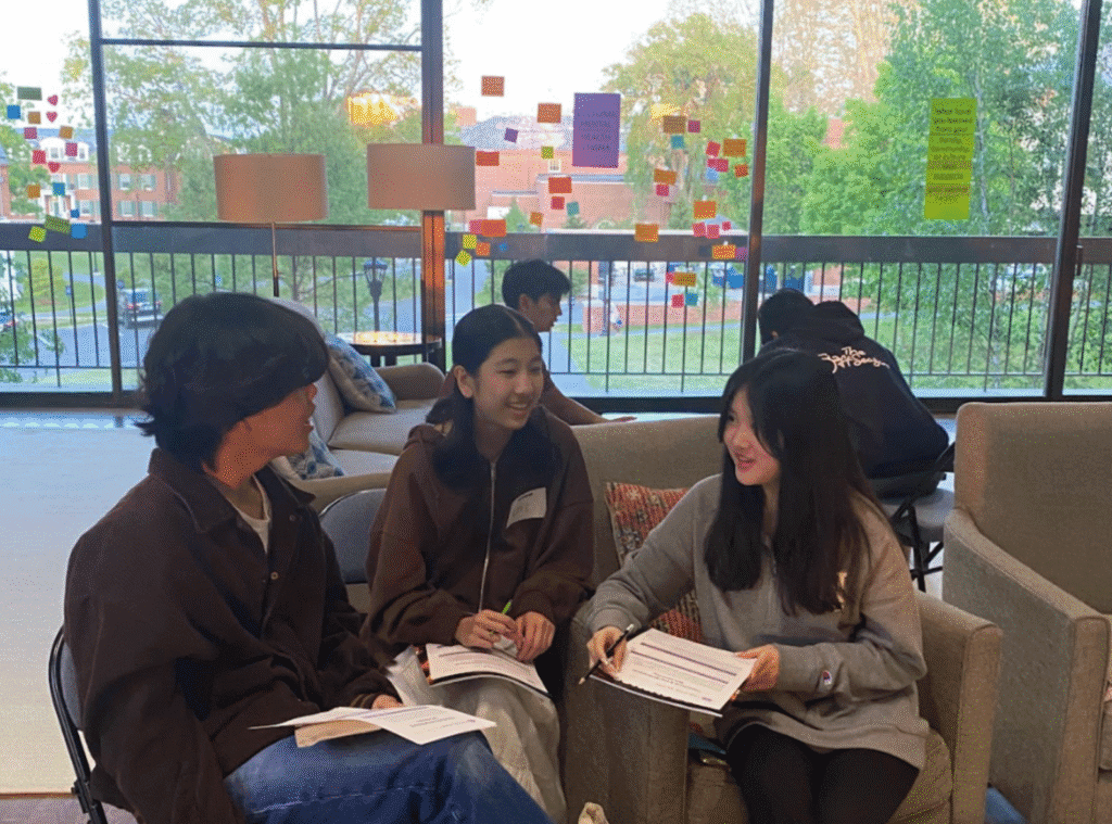 Three people sitting indoors on a sofa and chairs, engaged in conversation and writing on notepads, with a large window in the background displaying various colorful sticky notes.