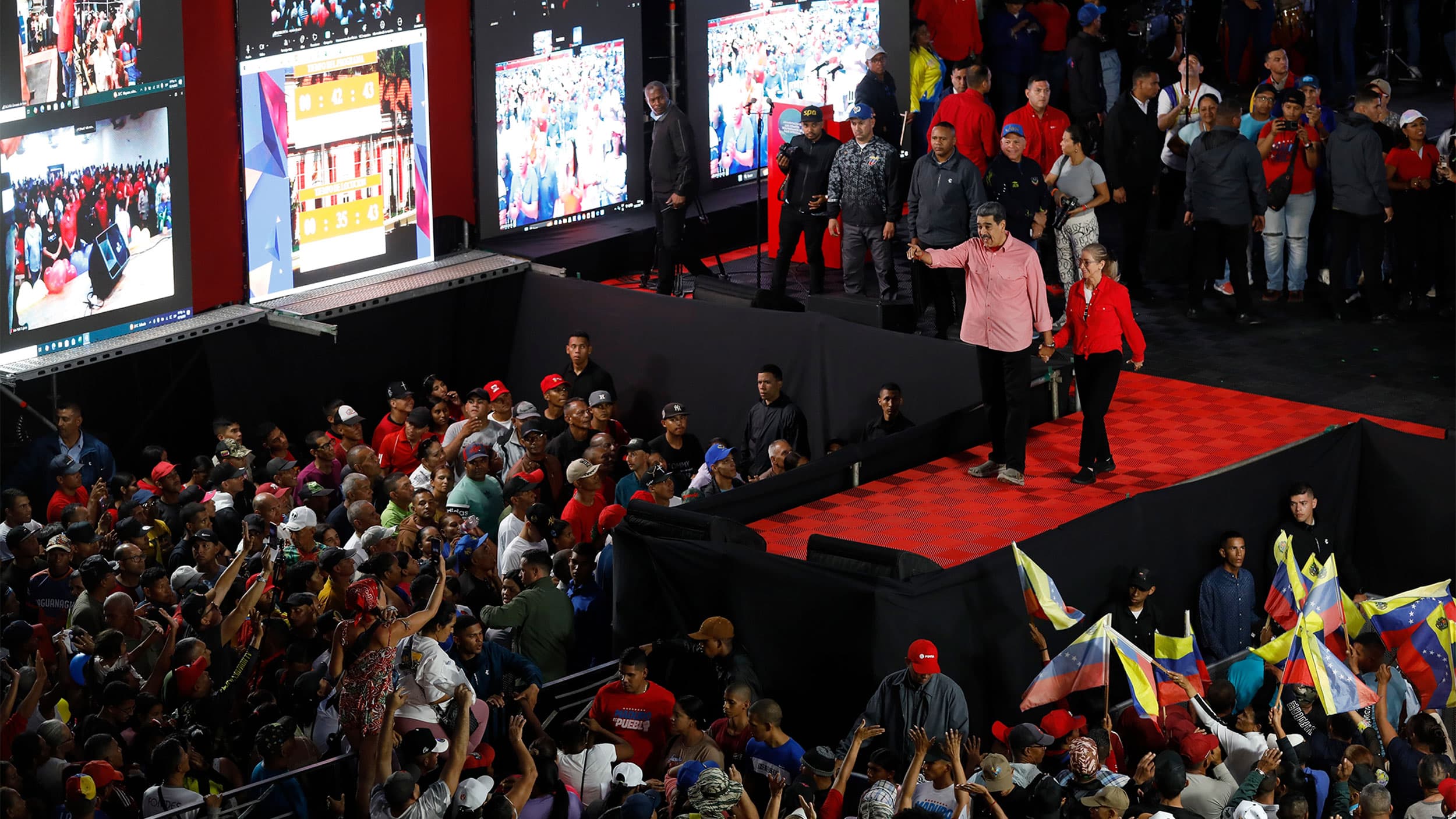A large crowd gathered at a political rally with prominent figures on stage, surrounded by many supporters waving flags and taking photos. Multiple large screens display images from the event.