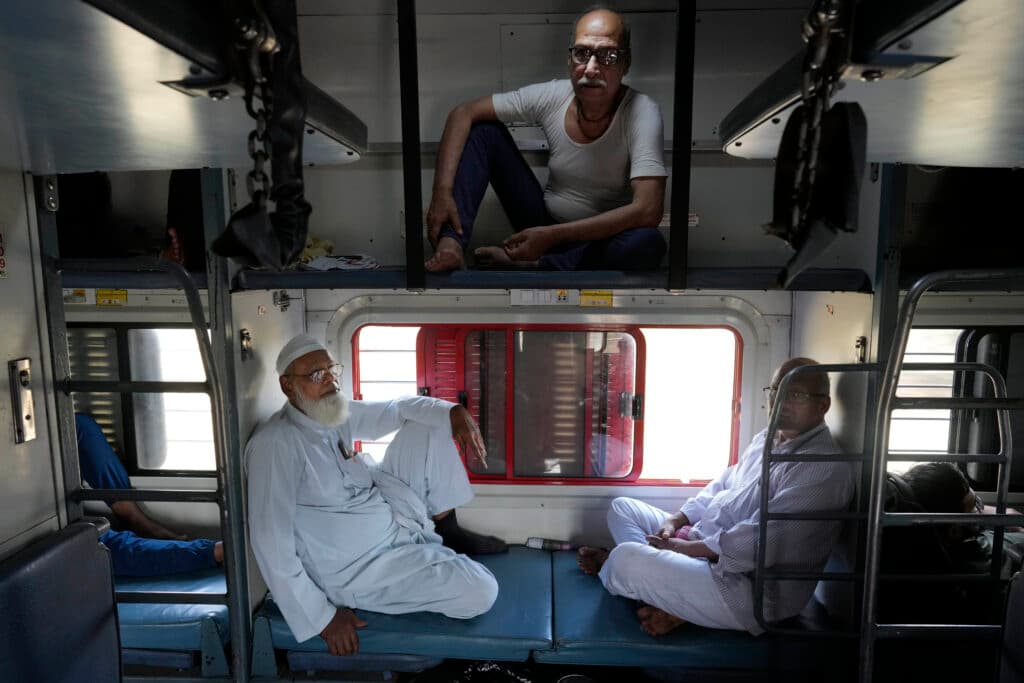 Passengers seated on bunk beds inside a train compartment, with one man sitting on an upper berth and two others on the lower berths, near a window with bars.