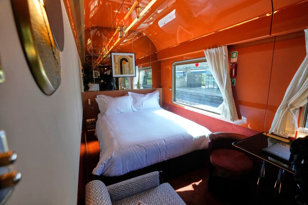 Interior of a luxurious train cabin with a plush white bed, orange walls, a large window with curtains, and a small seating area.