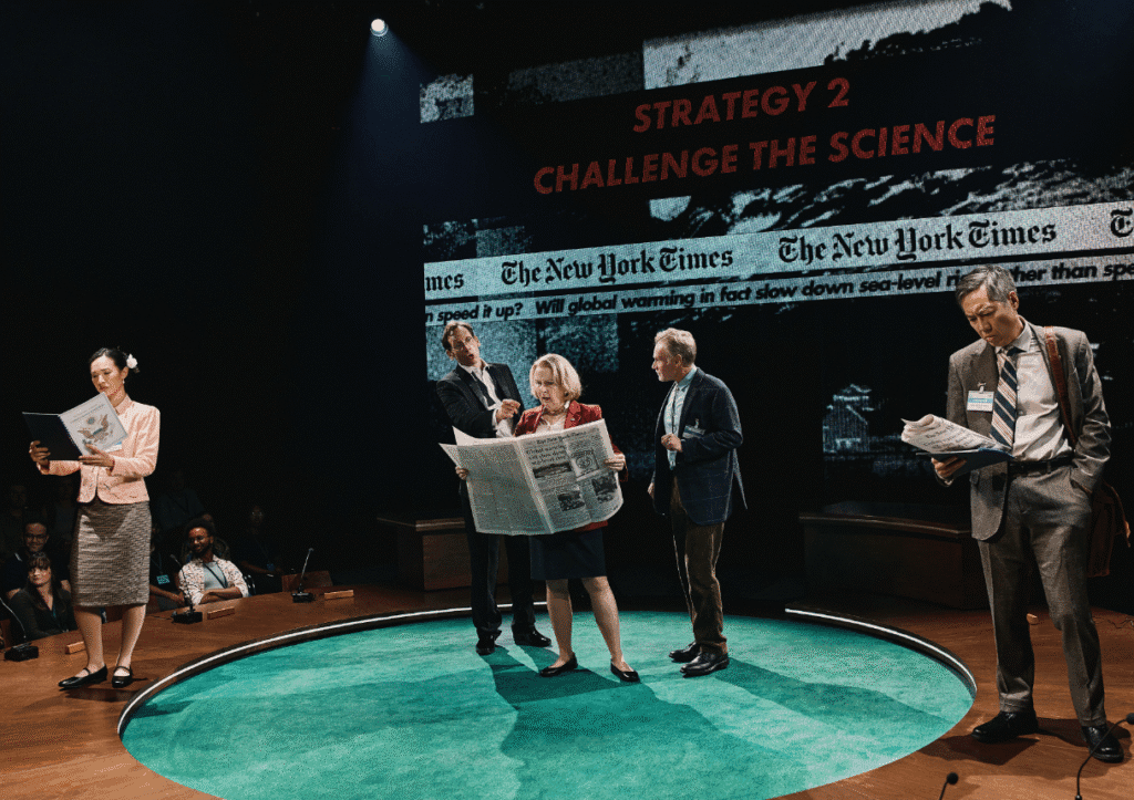 A group of actors perform on stage with a backdrop displaying headlines from The New York Times and the phrase "Strategy 2: Challenge the Science." One woman in the foreground is reading a large newspaper, while others examine documents, suggesting themes of media and climate change discussion.