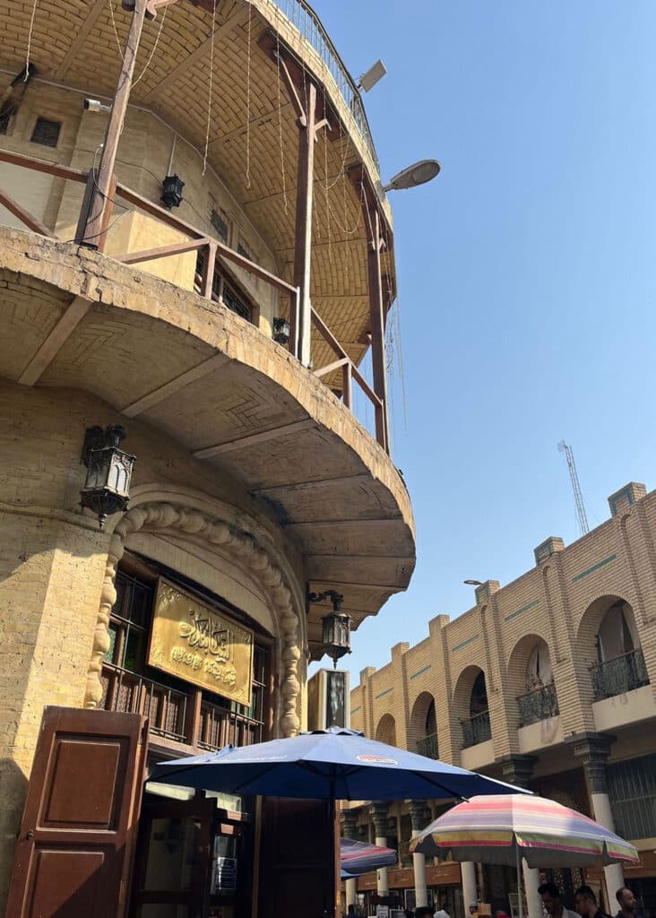 A street scene featuring a historic building with arched windows and decorative lanterns, and a modern building with umbrella-covered stalls below a clear blue sky.