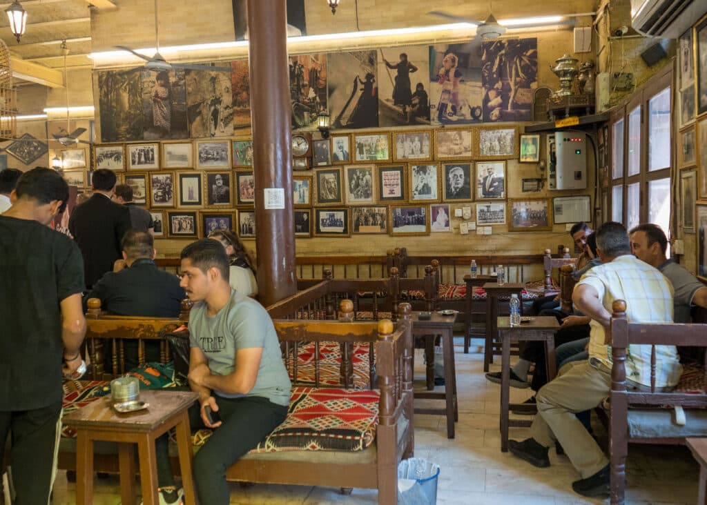 Interior of a busy, traditional café with people sitting on benches adorned with patterned cushions. The walls are covered with framed photos and memorabilia. A large wooden pillar stands in the center, and there are wooden tables and chairs throughout the space.