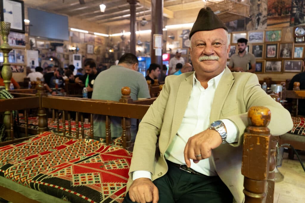 A man wearing a brown hat and beige jacket sits in a traditional cafe with patterned cushions, surrounded by people and pictures on the walls.