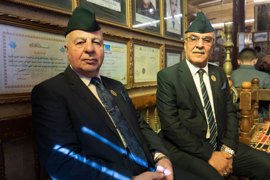 Two men in suits and caps sit on chairs in a room decorated with framed documents and photographs.