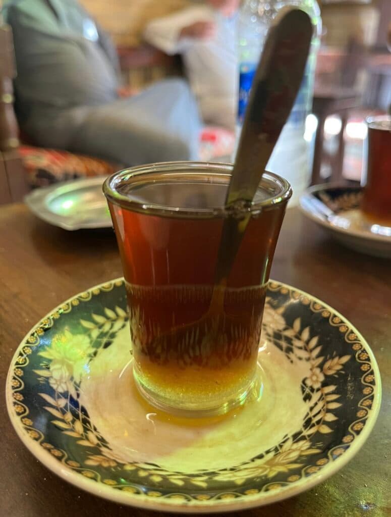 A glass of dark tea with a spoon inside, resting on an intricately patterned saucer, with a blurry background of people and a water bottle in a café setting.