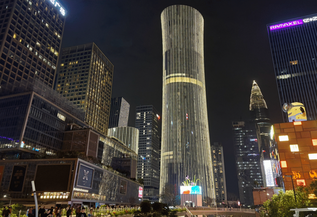 A cityscape at night featuring modern skyscrapers illuminated by lights. The tallest building is cylindrical with vertical stripes of light, surrounded by other high-rise buildings. A building with a peaked top is visible to the right. The street below is bustling with people and bright colorful signage.