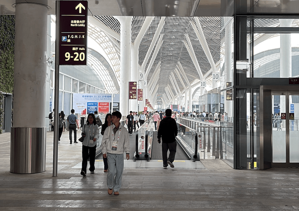 A spacious indoor hall with a high arched ceiling featuring skylights. People are walking in various directions, with some using a moving walkway. Signs above indicate directions to halls 9 to 20 in the North Lobby.