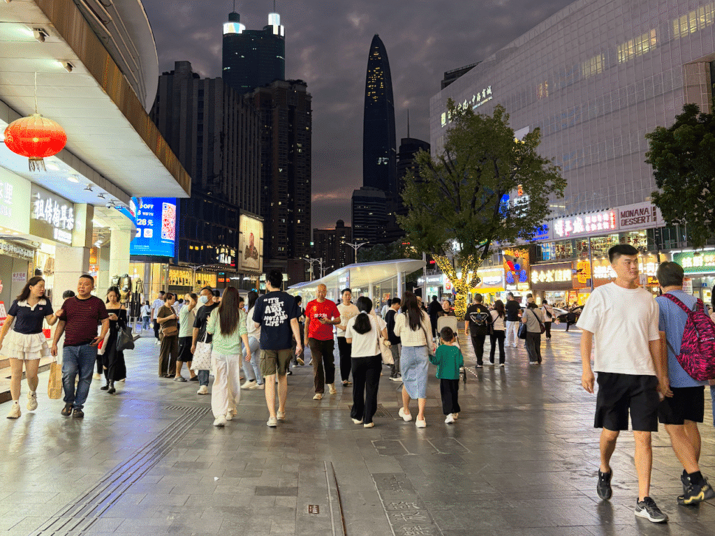 A bustling street scene at dusk in an urban shopping area, with people walking on a tiled pathway surrounded by lit storefronts and neon signs. Tall buildings rise in the background under a cloudy sky, and a tree decorated with lights stands on the side of the pathway.
