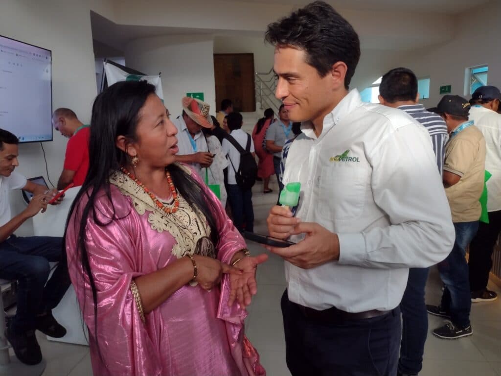A woman in a pink traditional dress is talking to a man in a white shirt with the 'Ecopetrol' logo, holding a green popsicle.