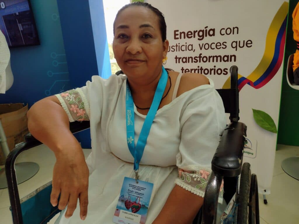 A woman seated in a wheelchair, wearing a white dress with floral embroidery on the sleeves, and a blue lanyard with a badge around her neck. A banner in the background features text related to energy and transformation, with a design resembling the Colombian flag.