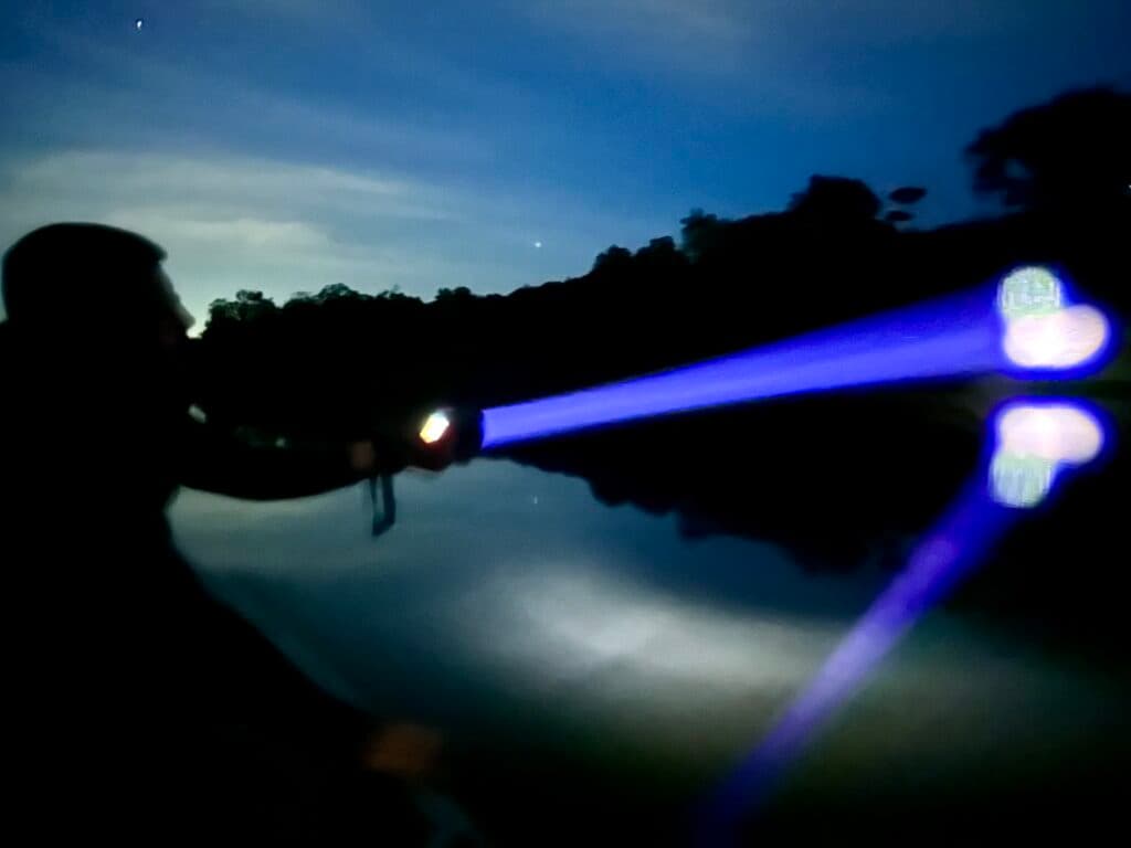A person using a blue flashlight near water at night, with the light reflecting off the water's surface.