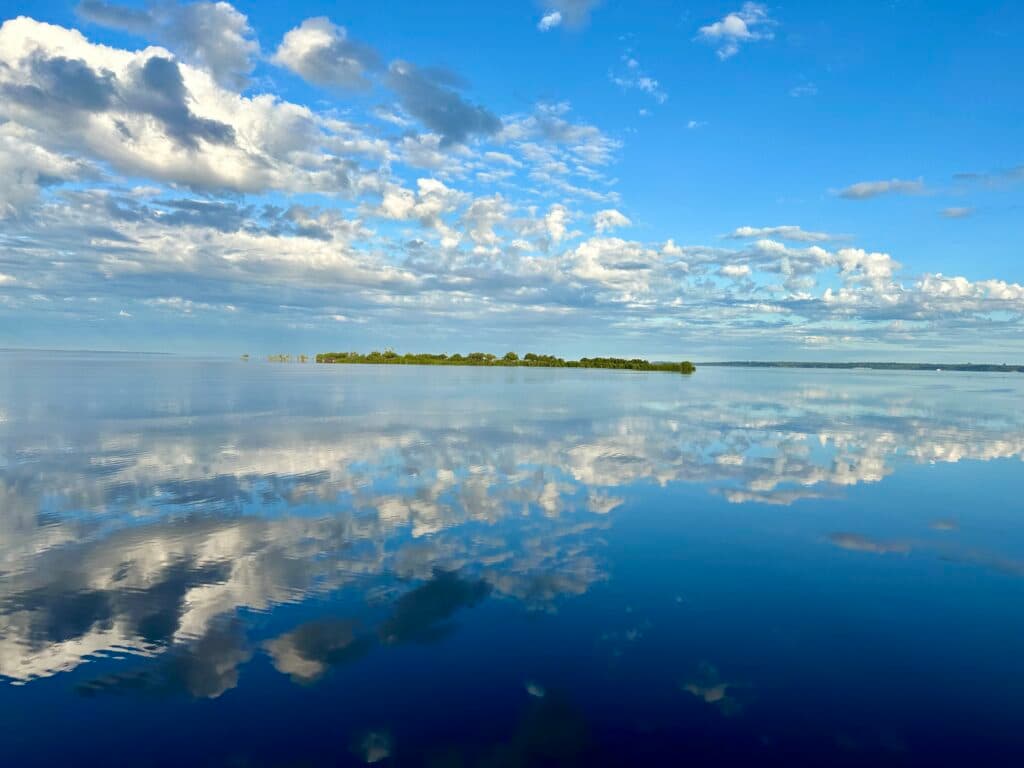 A calm lake reflecting a blue sky filled with white clouds and a distant green island on the horizon.