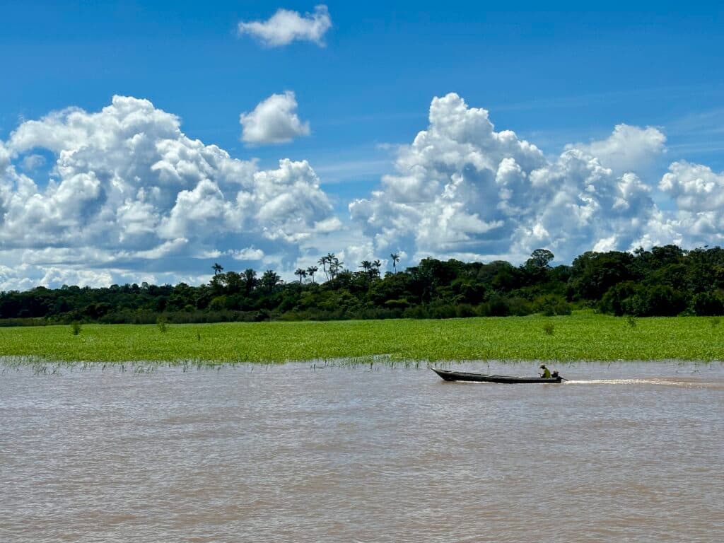 A small wooden boat with two people is moving across a brown river, with a lush green landscape and dense trees in the background under a blue sky filled with large, fluffy clouds.
