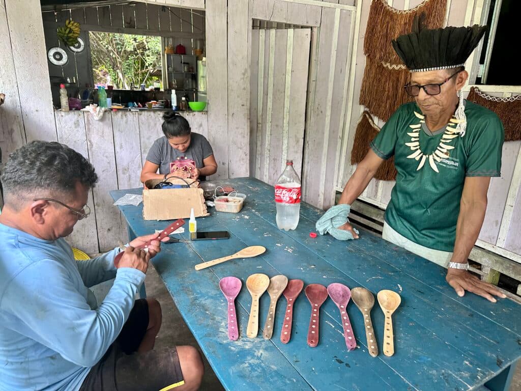 Three people in a rustic kitchen setting, with one person carving a wooden object, another using a mobile device, and a third observing from the side, wearing a traditional feathered headdress and a bone necklace, with wooden spoons displayed on the table.