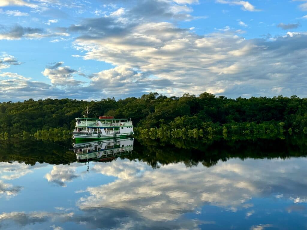 A green and white boat is reflected on a calm river with lush trees in the background under a partly cloudy sky.