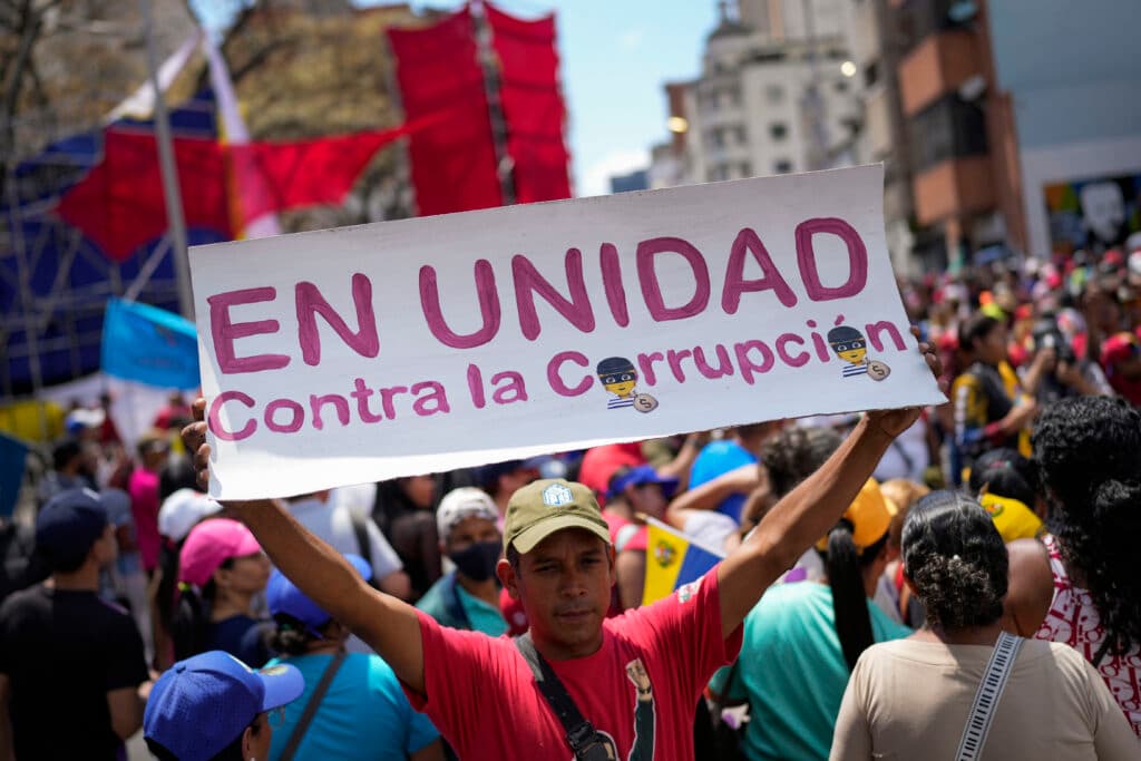 A man holds a sign reading "EN UNIDAD Contra la Corrupción," amidst a large crowd of people participating in a protest or rally.