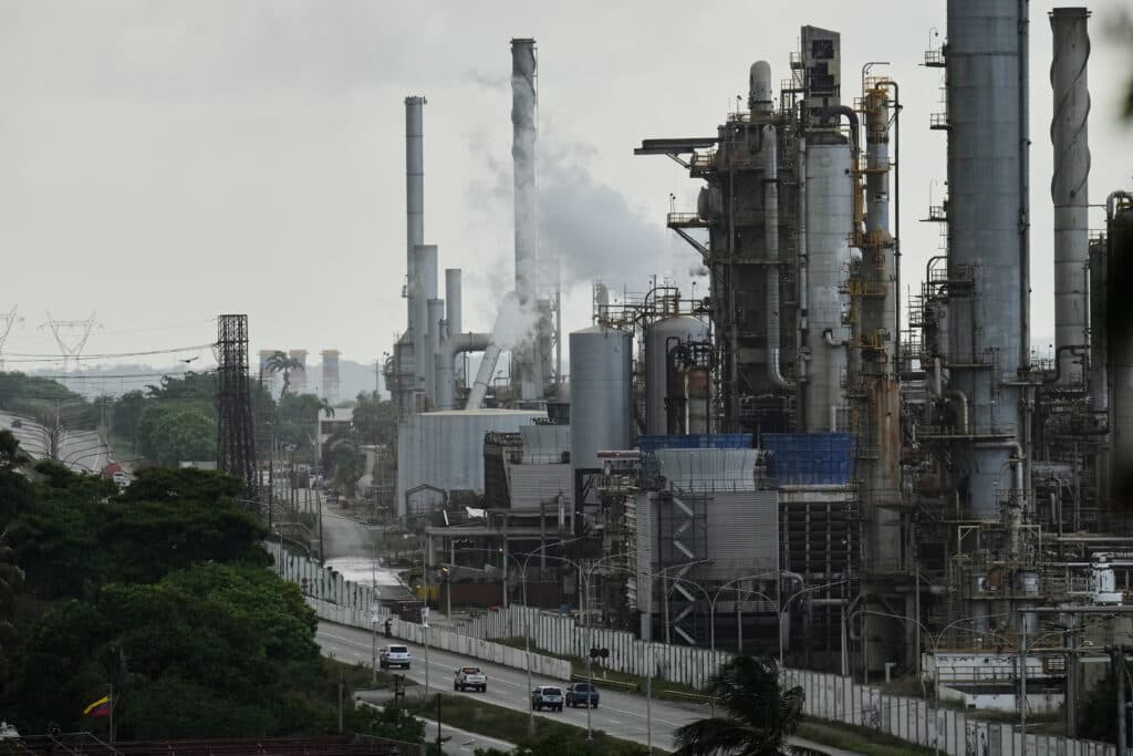 Industrial refinery complex with tall smokestacks emitting smoke, adjacent to a road with several vehicles, surrounded by trees and power lines under an overcast sky.