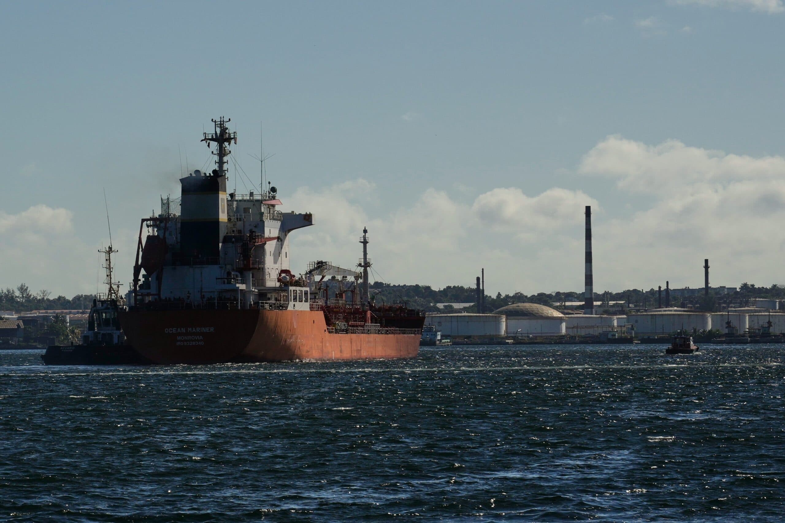 A large cargo ship with a red hull and white superstructure is navigating through water with an industrial facility and smokestacks visible in the background under a partly cloudy sky.