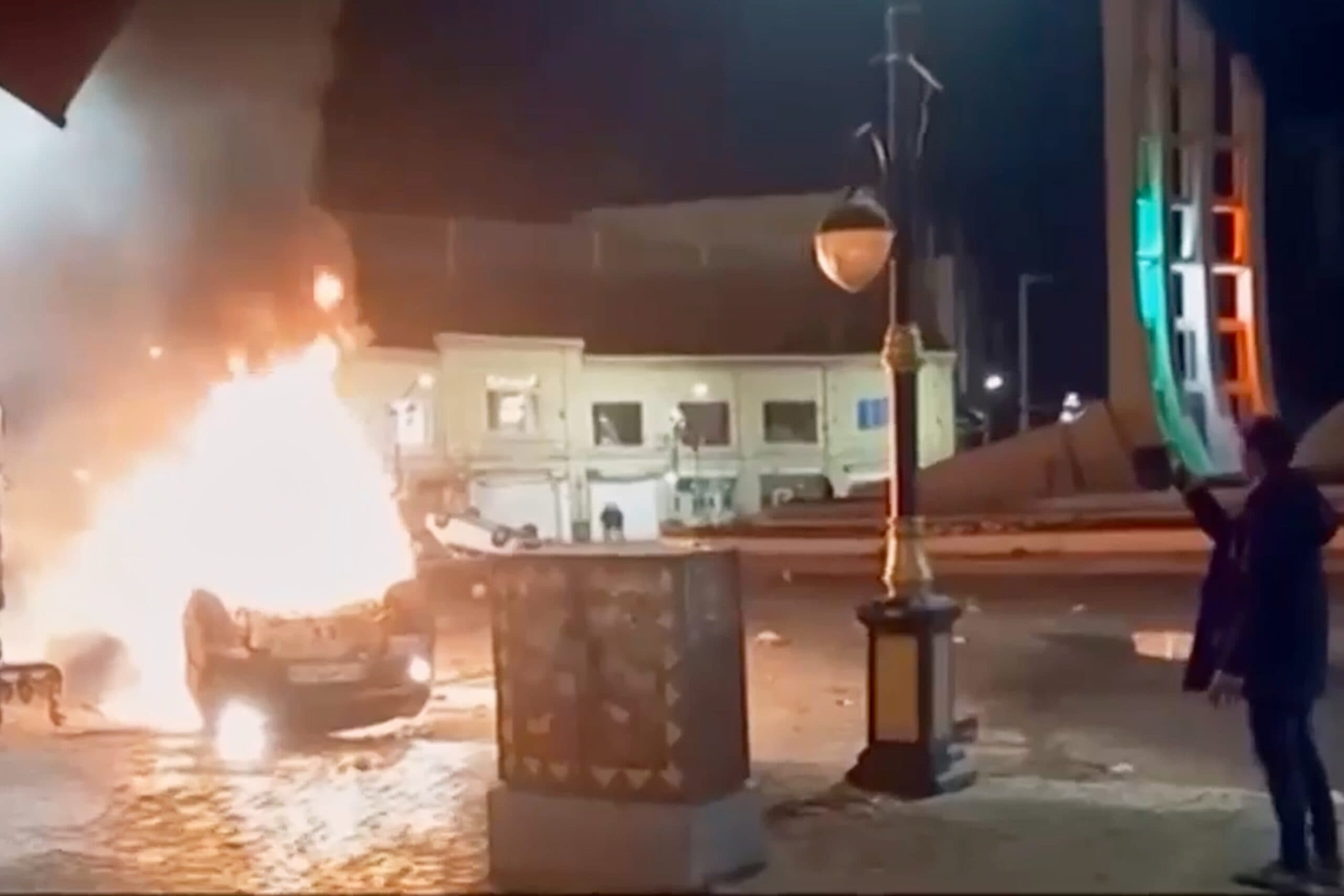 A car on fire in a public square at night, with flames and smoke rising into the air. A person stands nearby observing the scene, with buildings and a decorated structure illuminated in the background.