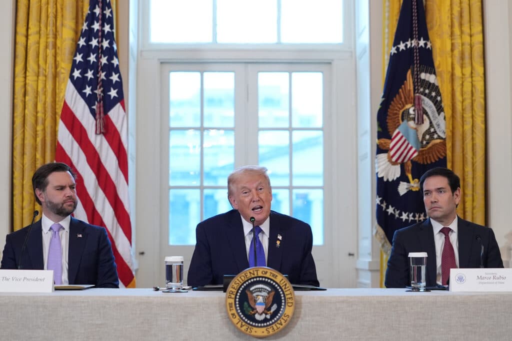 Three men seated at a table during a formal event, with one speaking into a microphone, under two American flags and the Presidential seal.