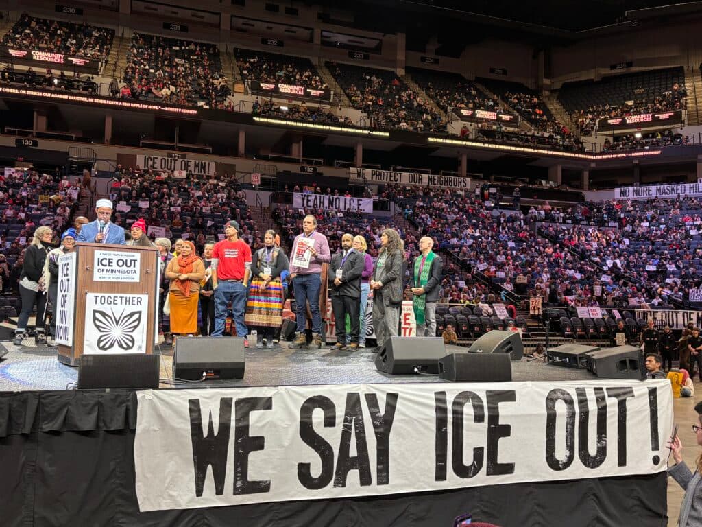 A group of people stand on a stage at an indoor arena, holding signs related to an "ICE Out of Minnesota" campaign. The banner on the podium reads "Together" with a butterfly symbol. The audience in the background holds various signs, and a large banner in the foreground states, "We Say ICE Out!"