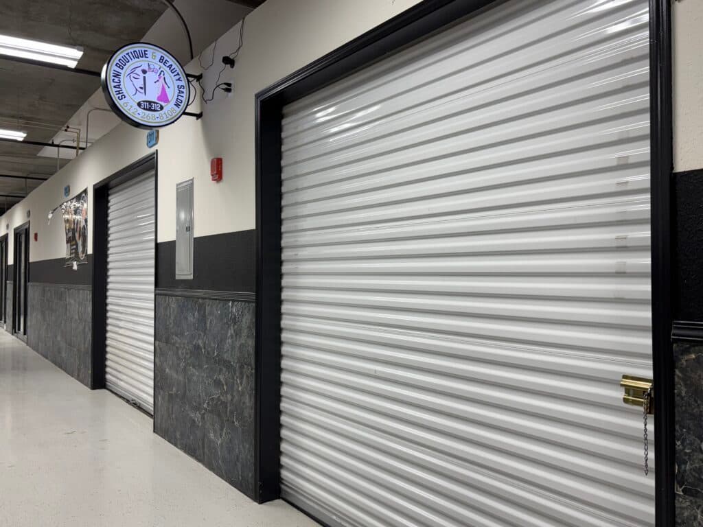 An indoor hallway with closed white, rolling metal shutters on multiple storefronts. A round sign for "Shacina Boutique & Beauty Salon" is mounted on the wall above one of the shops. The floor is polished and walls are partially covered with marbled panels.