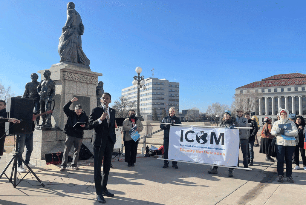 A speaker addressing a crowd at a rally beside a statue, with participants holding banners including one from the Interfaith Coalition on Immigration. The setting is a public plaza with a clear blue sky.