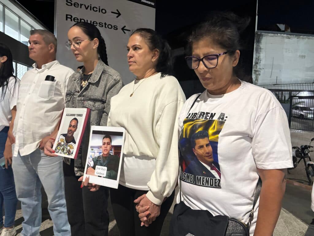 A group of people standing together, two in the center holding signs with photos and names "Alejandro Torres" and "José Ruiz Geleon." One person wears a t-shirt with a photo and text "Liberen a Rosmel Mendez." They appear to be in a vigil or protest setting, with a sign in the background reading "Servicio Repuestos."