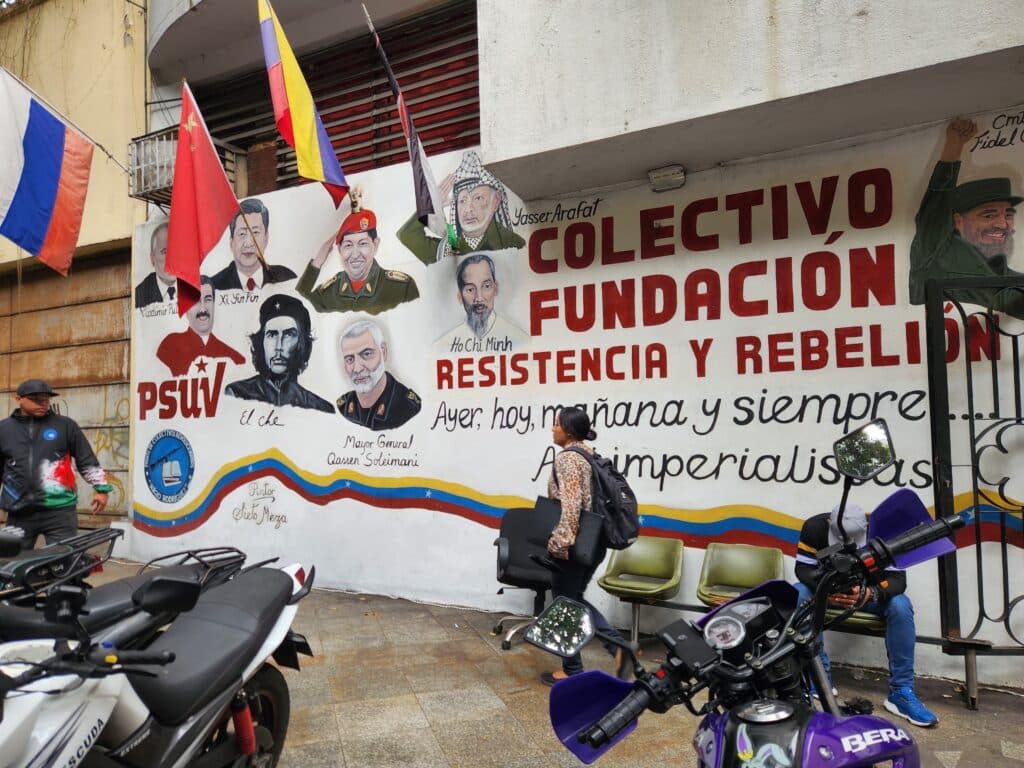 A mural featuring portraits of various political figures alongside flags and the words "Colectivo Fundación Resistencia y Rebelión" on a building wall, with motorcycles and pedestrians in the foreground.