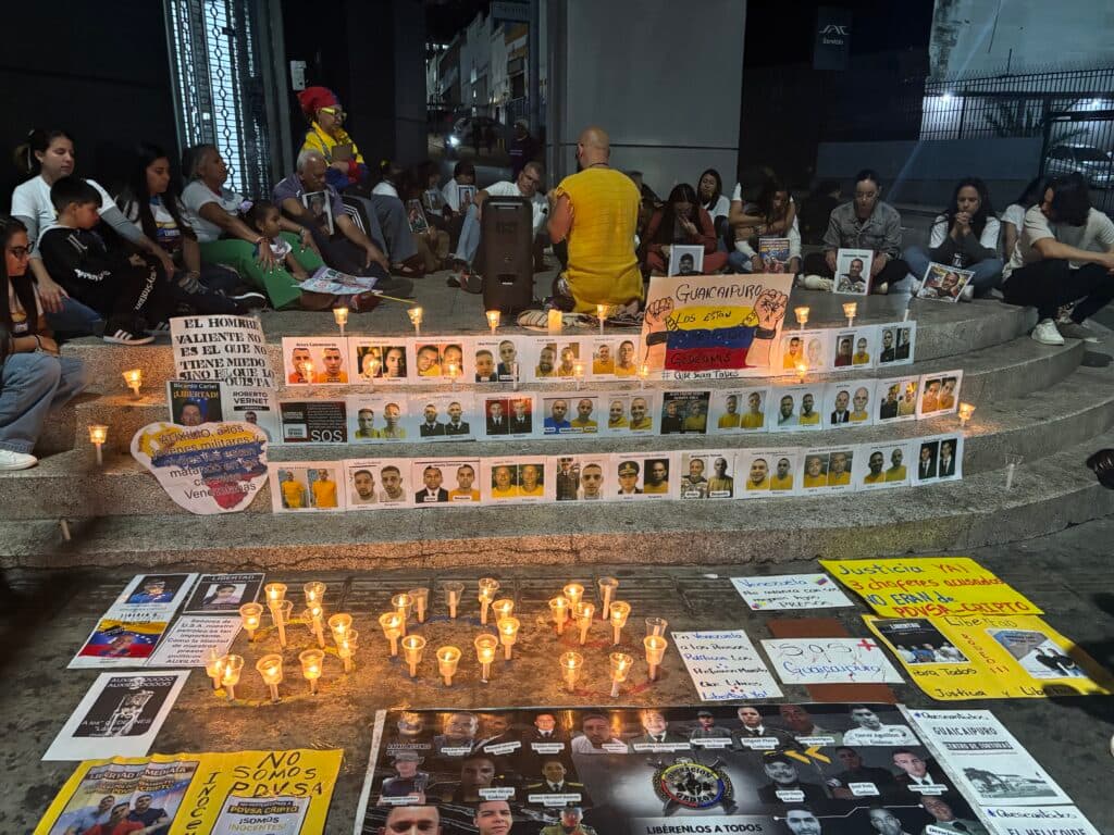 A nighttime protest scene featuring people sitting around lit candles on the steps, with posters and photos of individuals displayed. The gathering includes expressions of social and political demands, with slogans and messages related to justice and human rights. The atmosphere is somber and reflective.