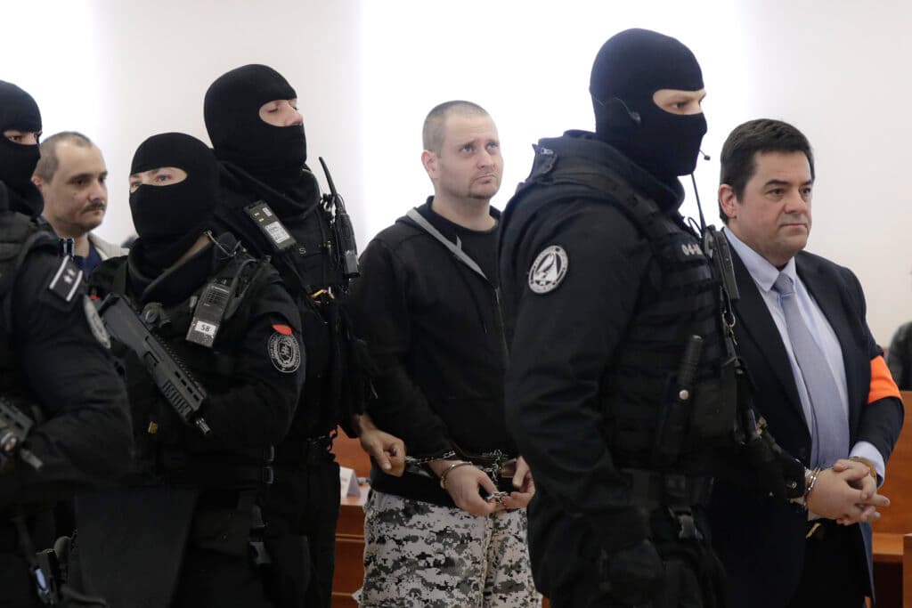 A group of masked security officers escort two handcuffed men in a formal setting. The officers are wearing black tactical gear and balaclavas, while the two men are dressed in civilian clothing.