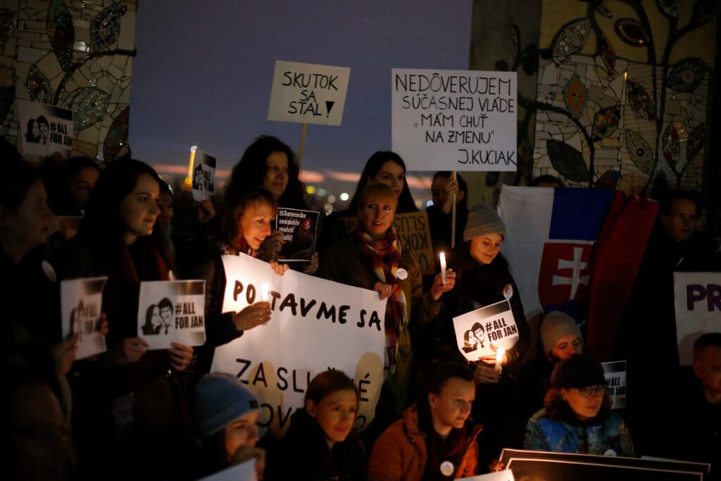 A group of people participating in a vigil at dusk, holding candles and signs with messages in Slovakian and "#AllForJan." A Slovak flag is partially visible in the background, and the setting has a mosaic-decorated structure.
