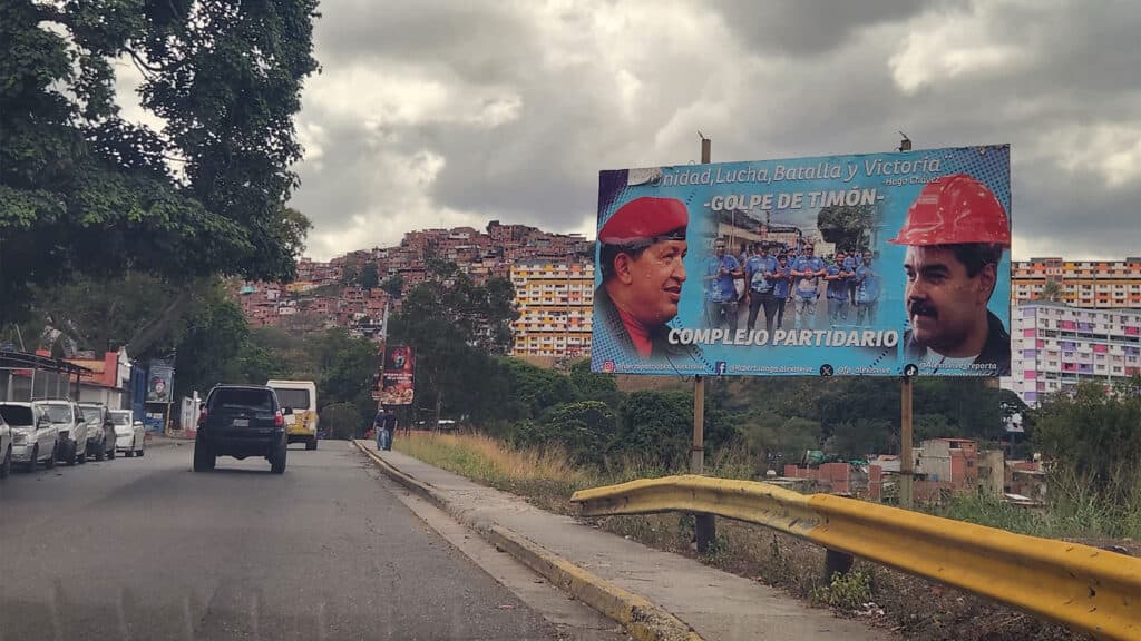 A roadside billboard in an urban area featuring two men, one wearing a red beret and the other a red hard hat. The billboard has text in Spanish and is set against a backdrop of a densely built hillside neighborhood. Several cars are parked along the road.