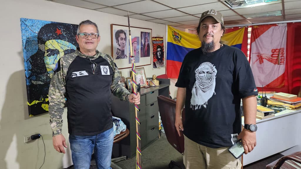 Two men standing in an office room, one holding a painted staff, with colorful wall art, flags, and framed pictures in the background.