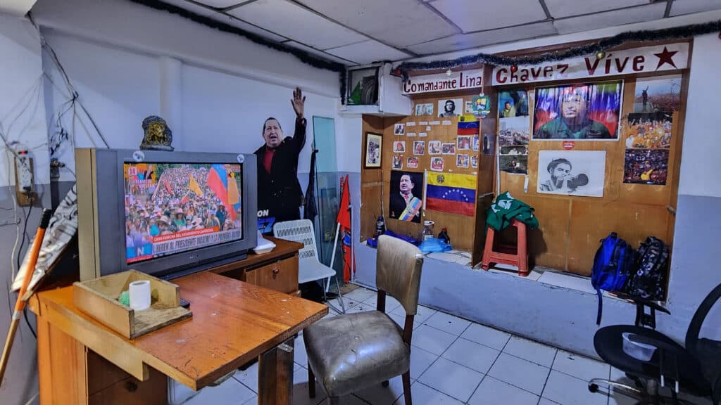 A room decorated with political memorabilia, including posters and a cardboard cutout of a well-known figure, with a Venezuelan flag displayed. A television is showing a broadcast with a crowd scene. Chairs and a table with miscellaneous items are present.