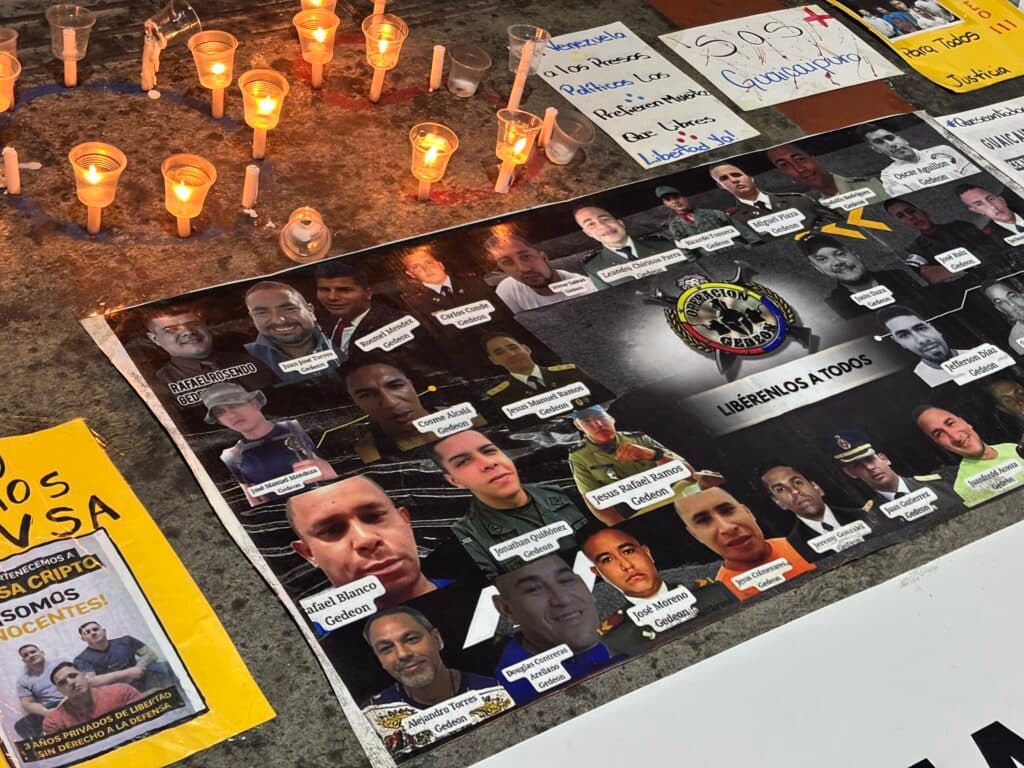 Close-up of a protest display featuring a collage of photos and names of individuals labeled as 'Gedeon,' candles in plastic cups, and handwritten signs advocating for freedom and justice in both Spanish and English.