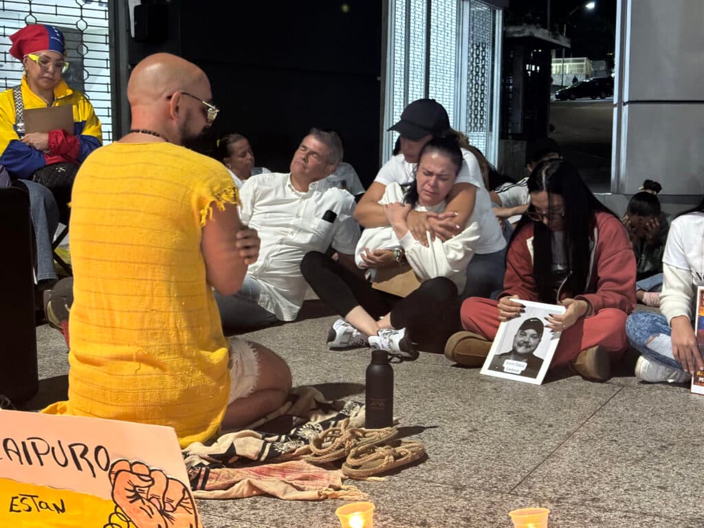 A group of people sit solemnly on the ground in front of a building, some holding photos, during a nighttime vigil. Candles are lit in the foreground, and a person in a yellow outfit sits with their back to the camera. One person is emotional, being comforted by another.
