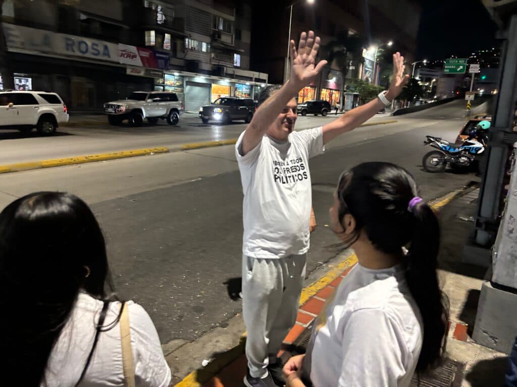 A person wearing a white shirt with text, standing on a sidewalk at night, raising their arms and appearing to greet or wave at oncoming traffic. Two people with long hair, also in white shirts, are facing the person with uplifted arms. Several vehicles are visible on the road, and it's a city setting with storefronts and streetlights illuminated.