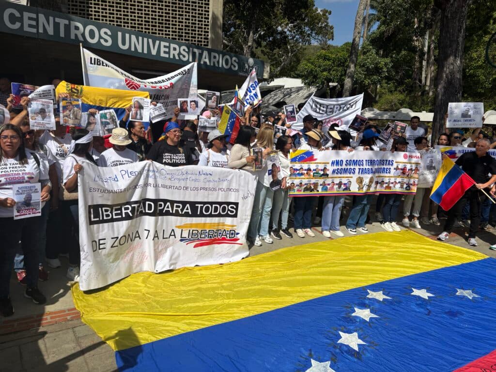 A group of people standing in front of a university building holding banners and signs advocating for the release of political prisoners in Venezuela. A large Venezuelan flag is displayed on the ground in front of them. The banners include phrases like "Libertad para todos" and "#No somos terroristas #Somos trabajadores," and feature images of individuals they are campaigning for.