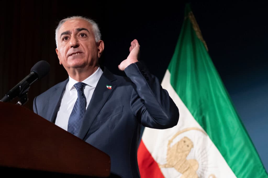 A man in a suit speaks at a podium with a microphone, gesturing with one hand. Behind him is a flag with a lion and sun emblem on green, white, and red stripes.