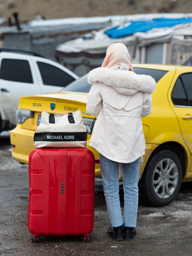 A person wearing a white fur-lined coat and jeans stands with a red suitcase and a Michael Kors bag near a yellow taxi labeled "Iran Turkiye."