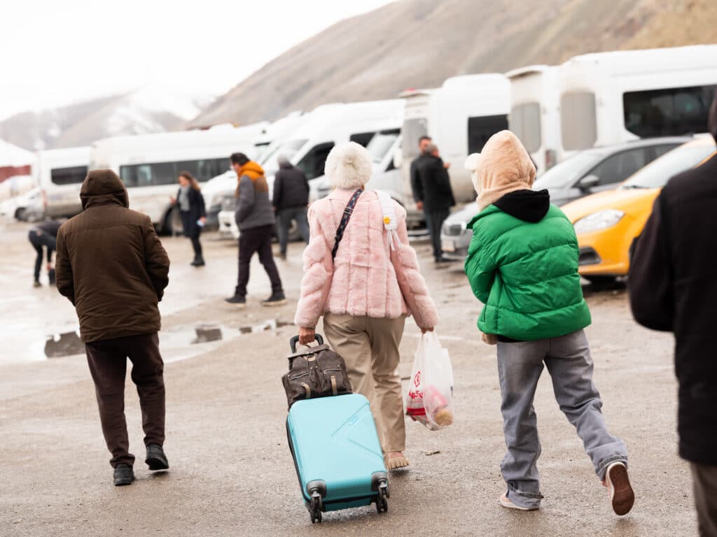 People in winter clothing walking in a parking area with white vans and other vehicles, carrying a turquoise suitcase and shopping bag.