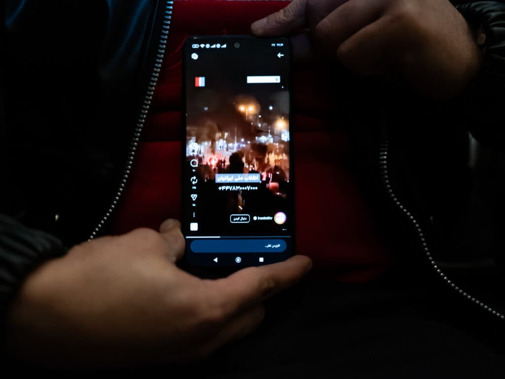 A person holding a smartphone displaying a video with Arabic text overlay, captured in dim lighting.