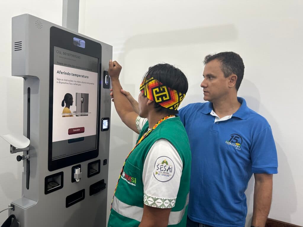 A man in a traditional headband and green vest is using a touchscreen machine to measure his temperature, while another man in a blue polo shirt observes beside him.