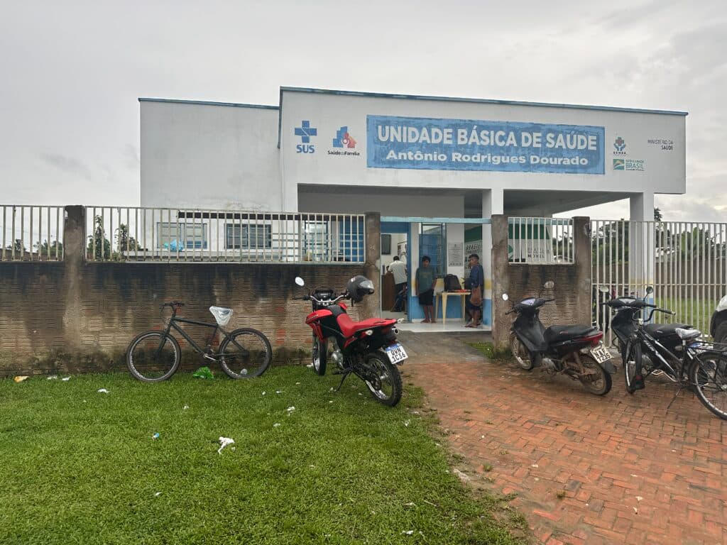 A health clinic building labeled "Unidade Básica de Saúde Antônio Rodrigues Dourado" with motorcycles and a bicycle parked in front, and people standing near the entrance.