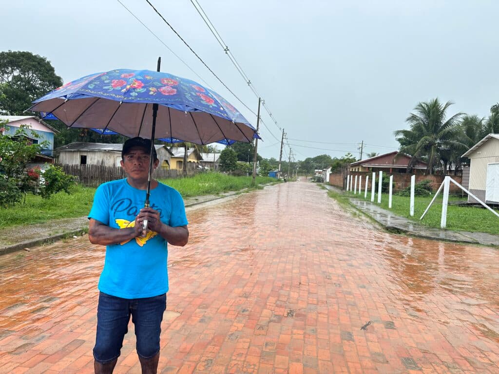 A man stands on a wet brick road holding an umbrella while wearing a blue T-shirt and jeans. The street is lined with houses and some palm trees, and the sky is overcast with rain.
