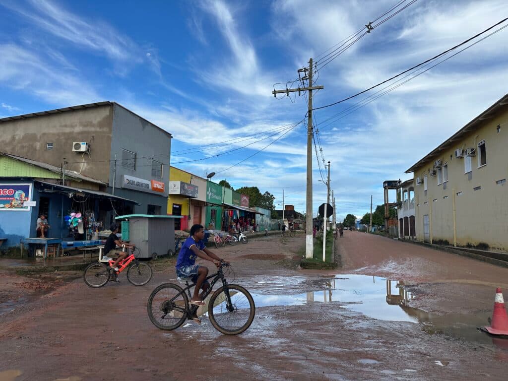 Two people riding bicycles on a muddy road with puddles in a small town, with shops and buildings lining one side of the street and a clear blue sky above.