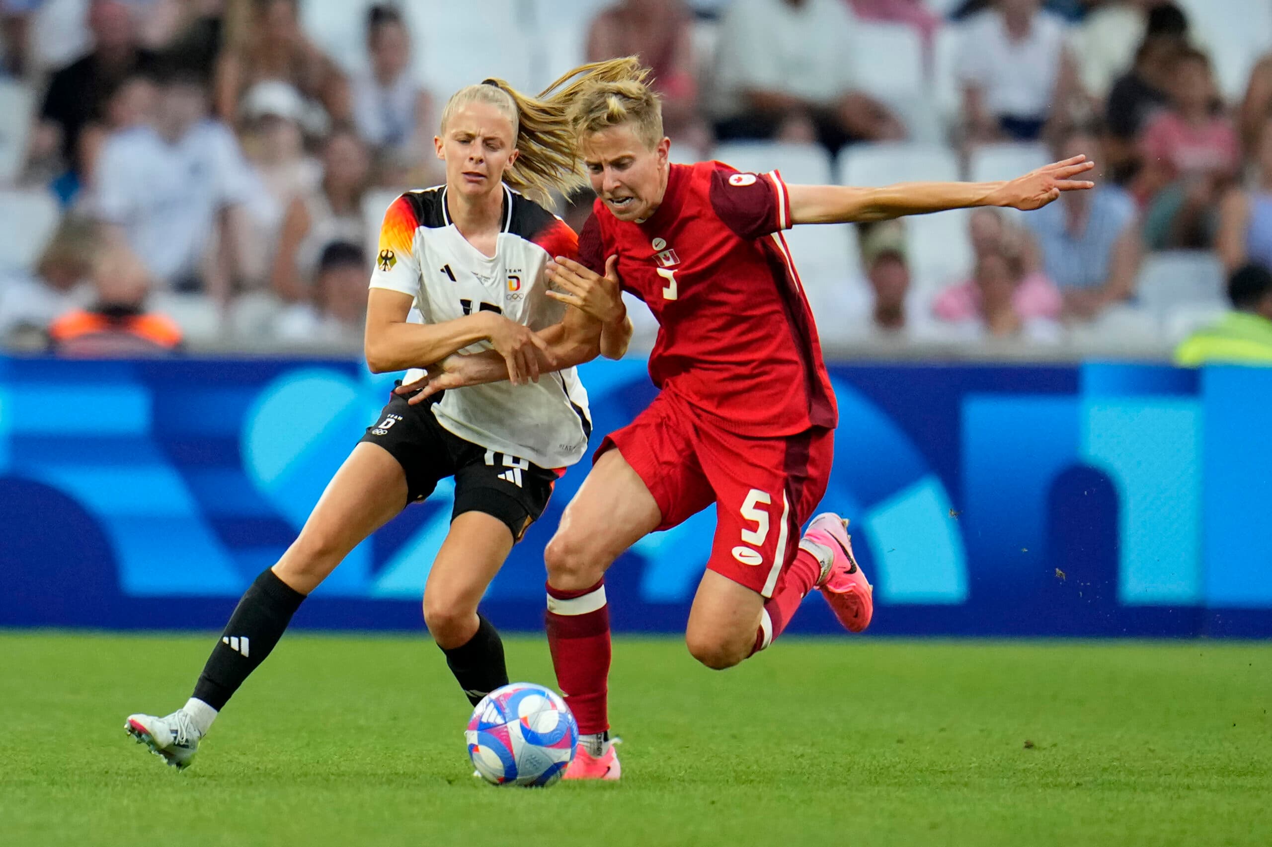 Two female soccer players competing for the ball during a match; one in a white jersey with black shorts, and the other in a red jersey and shorts. The scene takes place in a stadium with an audience in the background.